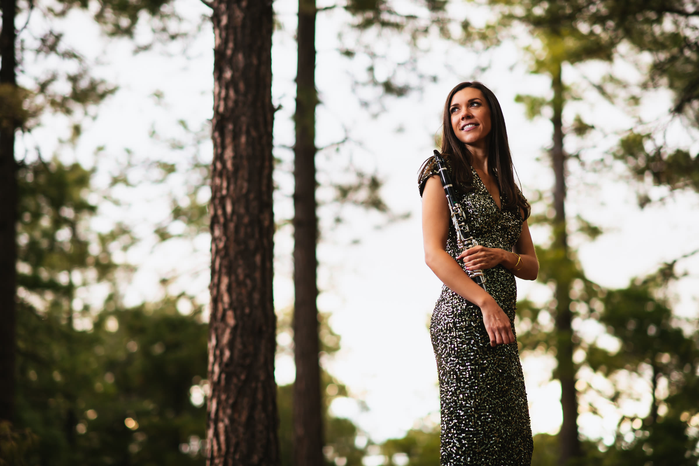 Jackie Glazier standing in a pine forest in a sequined gown, holding her clarinet